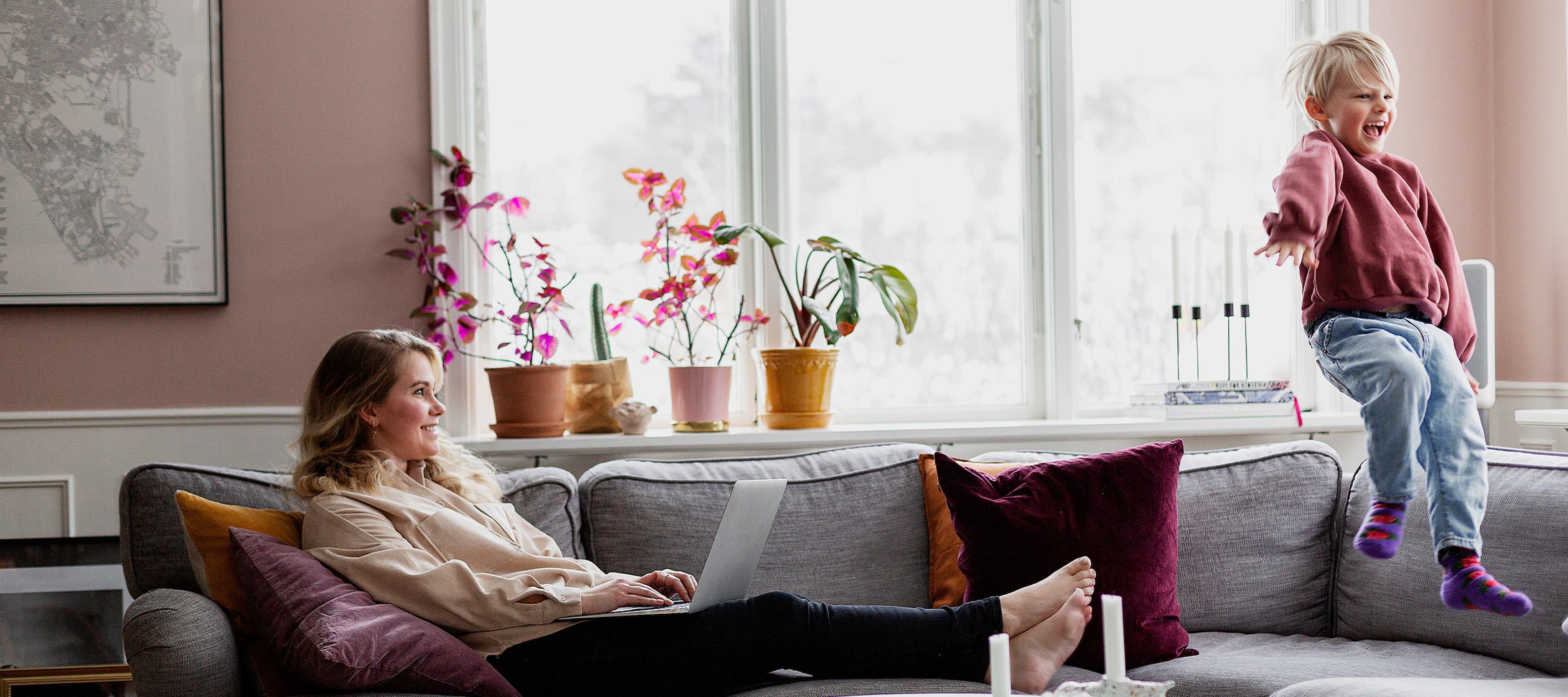 Kvinna sitter i soffan och tittar på ett barn som hoppar i vardagsrummet.