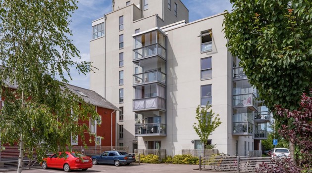 Flerbostadshus med ljus fasad och inglasade balkonger, sett från en parkeringsplats med träd i förgrunden.