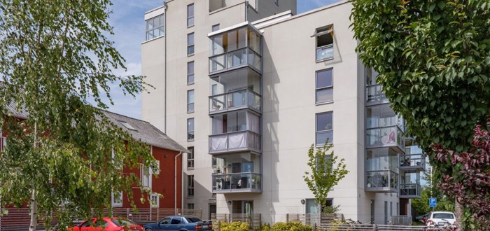 Flerbostadshus med ljus fasad och inglasade balkonger, sett från en parkeringsplats med träd i förgrunden.